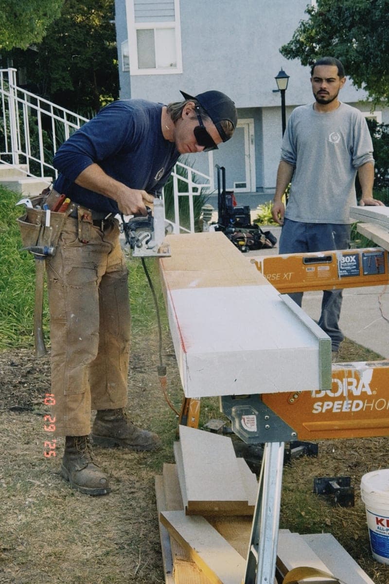 A carpenter running a saw along a long beam laid across sawhorses in a front yard, a second man watching the cut.