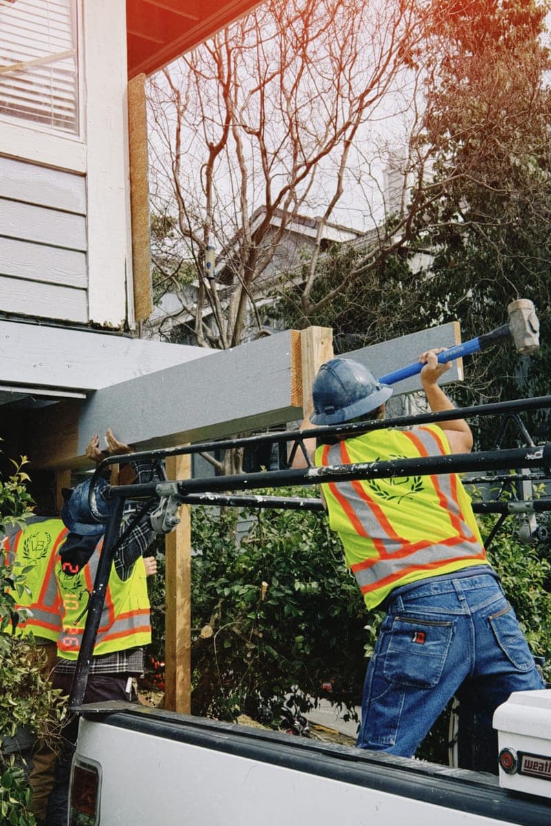 Three workers in high-visibility vests and hard hats walking a long beam into place above a porch, lifting it clear of a pickup bed.