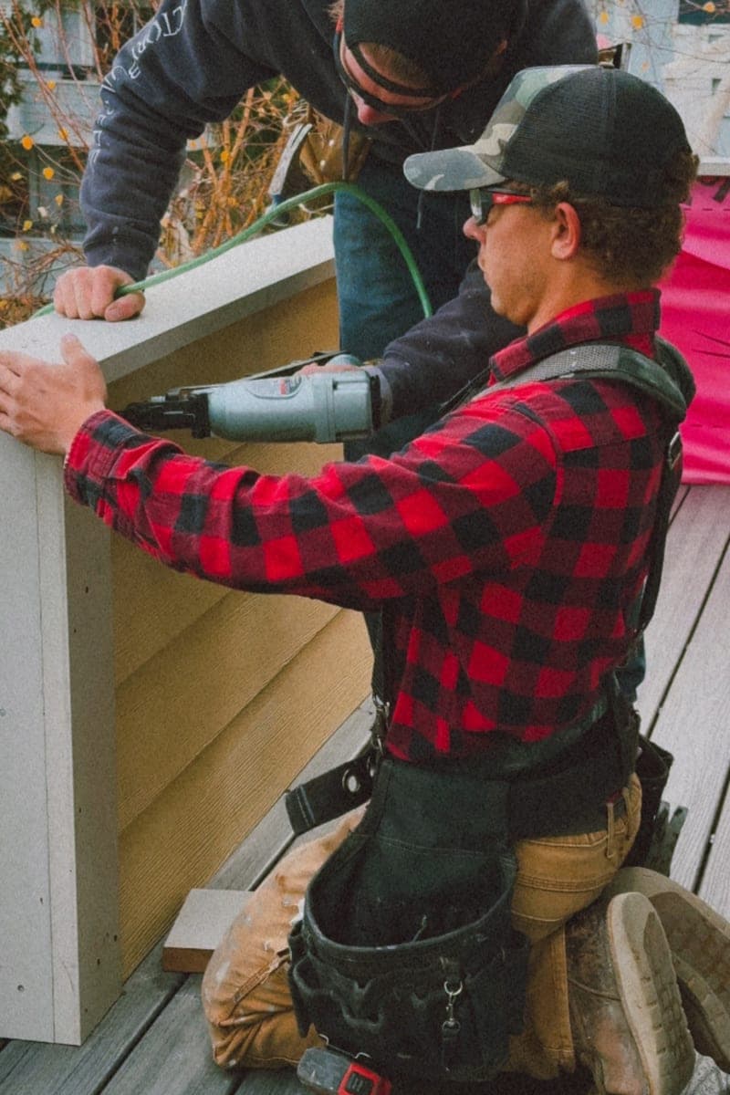 Two carpenters on a deck fitting a length of trim, one kneeling with a nail gun while the other holds the board level.