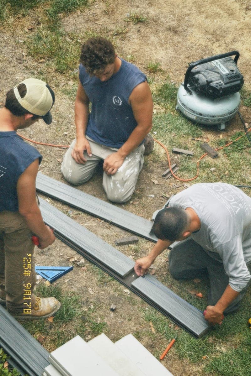 Three workers kneeling on the grass around a run of grey composite decking boards, an air compressor standing behind them.