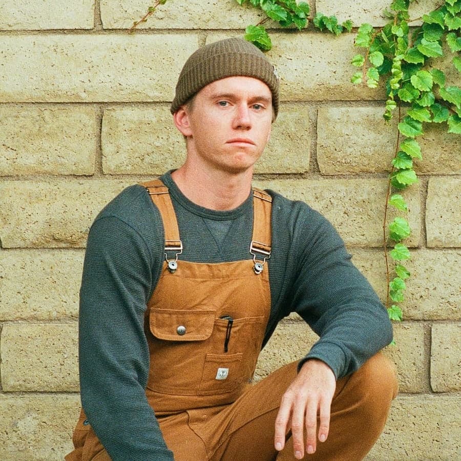 The person who built ClearBid, in work overalls and a beanie, sitting against a block wall with ivy growing down it.