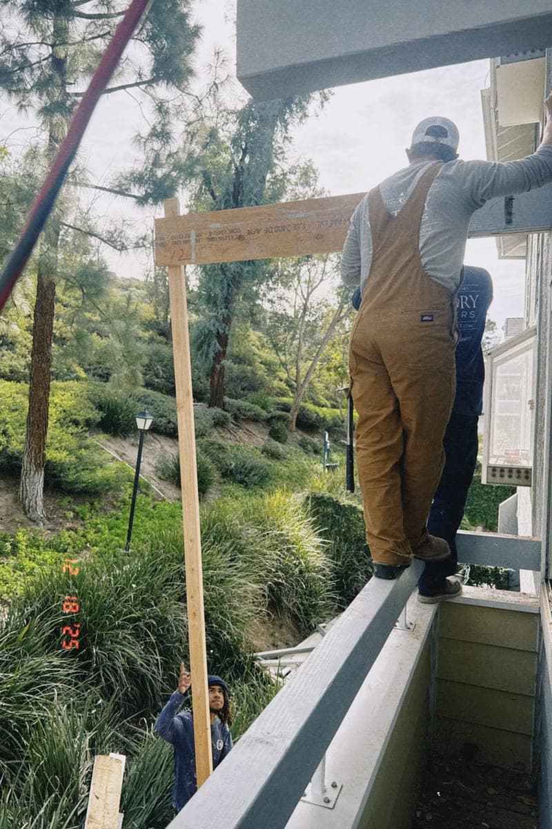 A worker in overalls standing up on a deck beam at height, a wooded hillside dropping away behind him.
