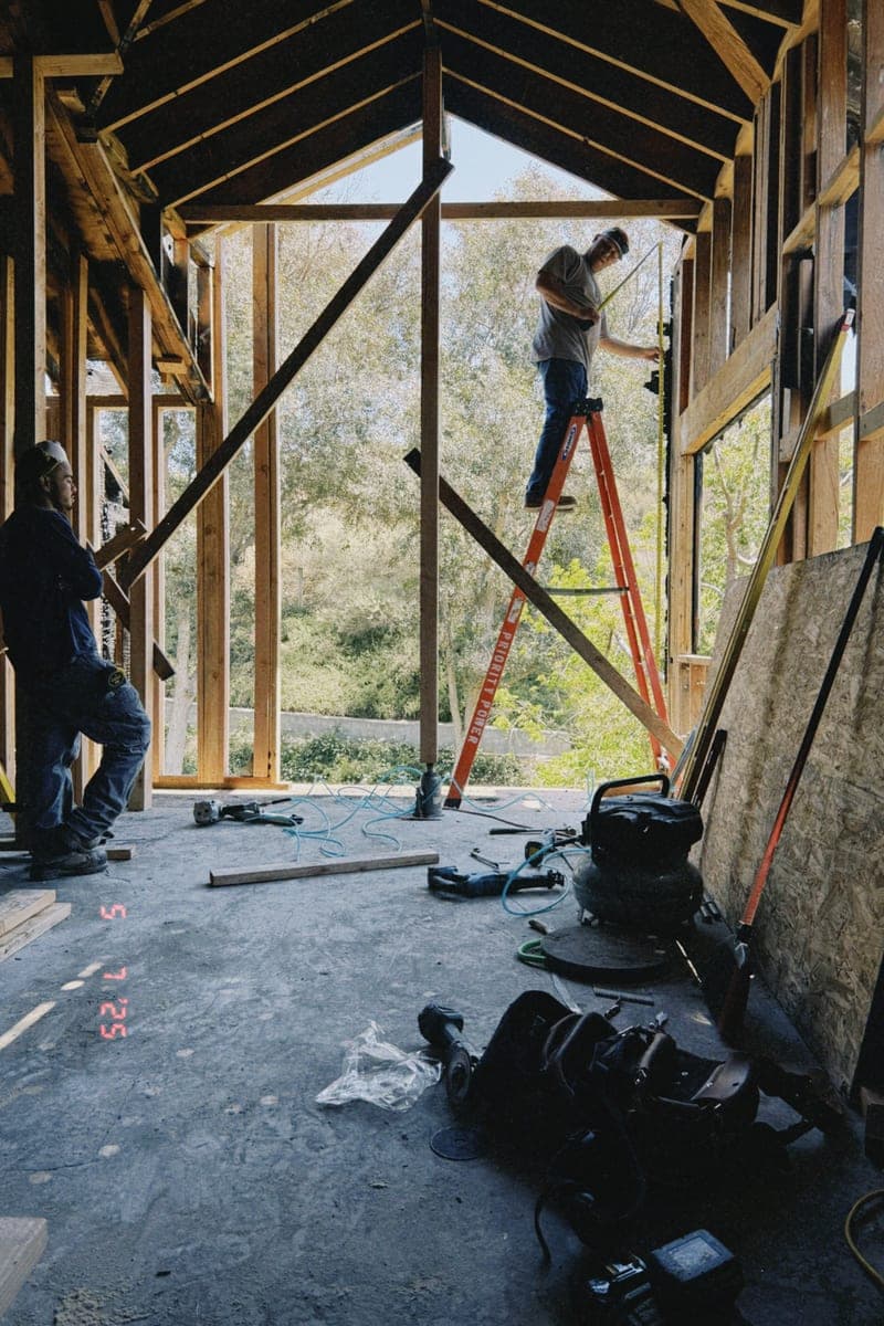 A building stripped back to its framing, with a worker on a ladder in a framed opening.