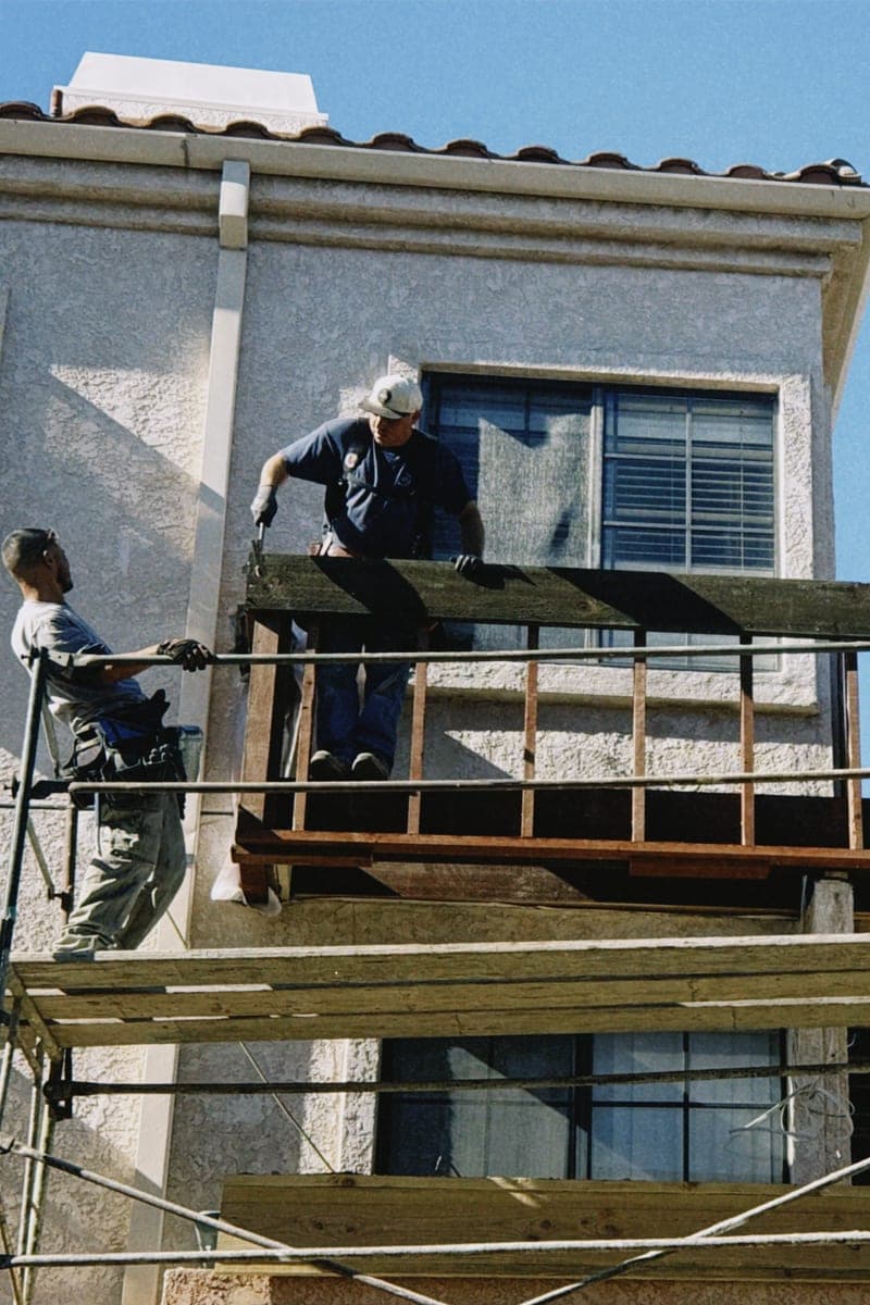 Two workers on scaffolding against the stucco upper storey of a house, building out a deck frame under a clear sky.