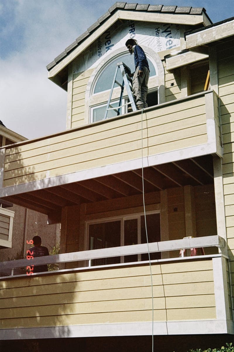 A worker up a ladder at a second-storey dormer window, finishing the siding above a newly built balcony.